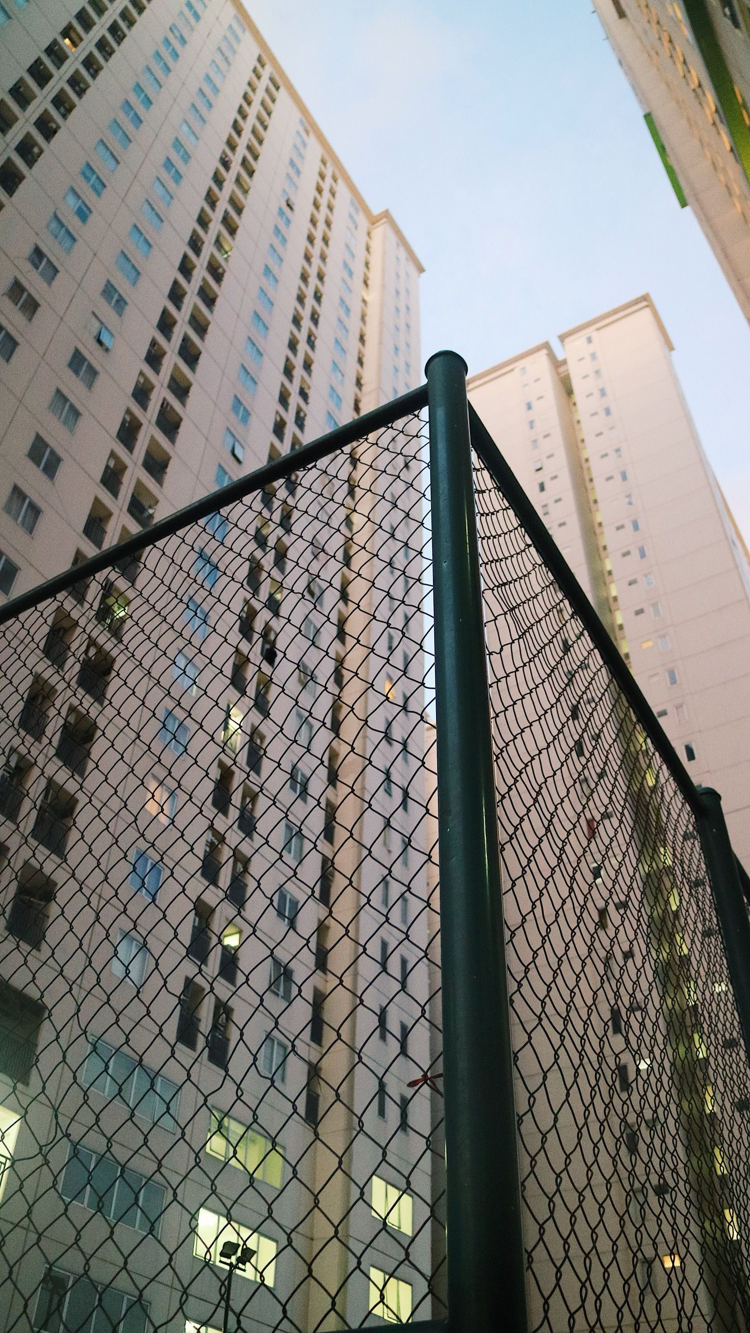 Low angle shot of tall buildings framed by a chain-link fence in an urban setting.